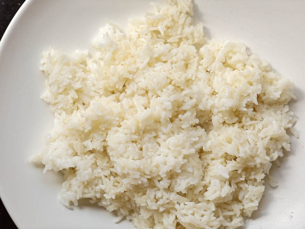 A white plate filled with cooked white rice, with individual grains visible and slightly fluffy in texture. The plate is positioned on a dark surface.